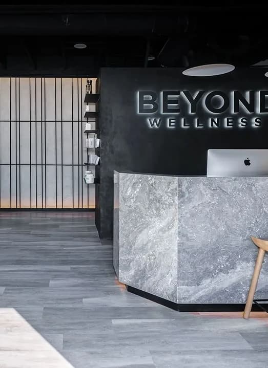 The Beyond Wellness reception: a grey marble desk under the backlit wordmark, black slat screens and a pale wood floor.
