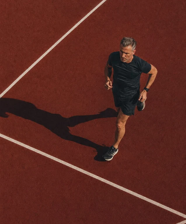 A man running on a red athletics track, seen from above with a long shadow.