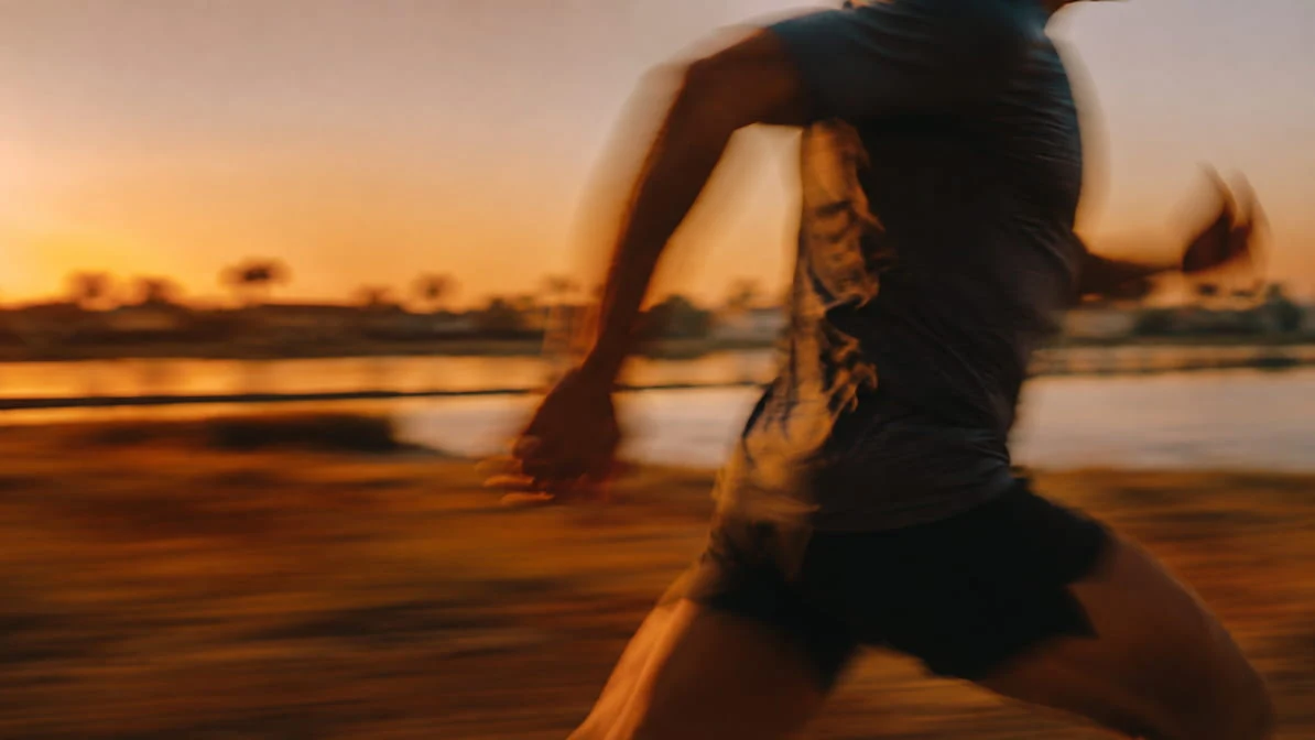 A man running at speed along a waterfront at sunset, caught with motion blur.