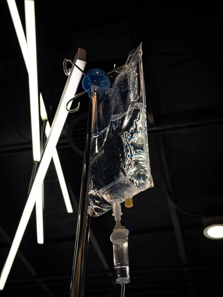 An IV bag hanging from its stand, seen from below against a dark ceiling.